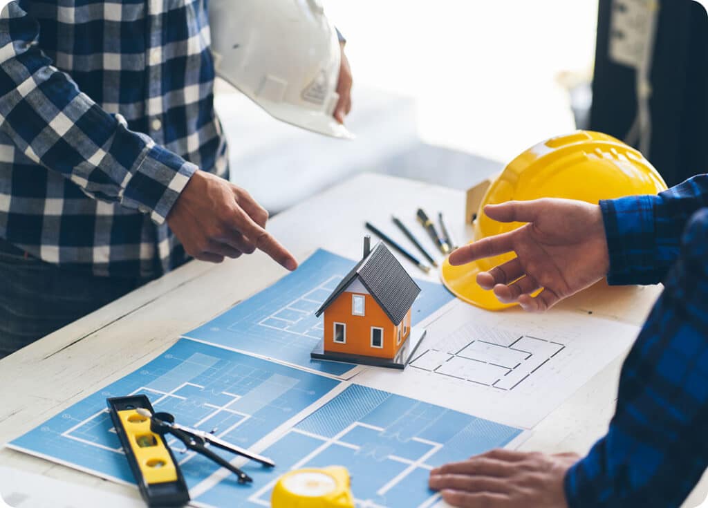 Close-up of two engineers discussing structural blueprints with a 3D house model and yellow hard hats on a project table.