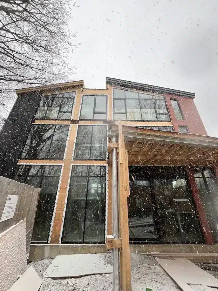 Wide-angle exterior view of a multi-storey residential construction site in the snow, showing stacks of lumber in the foreground and a modern house with red brick and black cladding.