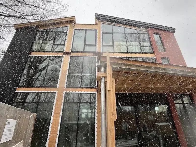 Wide-angle exterior view of a multi-storey residential construction site in the snow, showing stacks of lumber in the foreground and a modern house with red brick and black cladding.