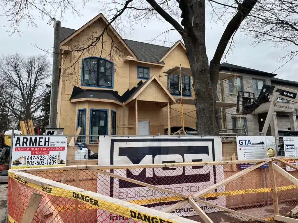 Mid-construction exterior view of the custom residential building, highlighting plywood wall sheathing, a lower curved bay window frame section, and a corporate builder banner on the front chain-link fence.