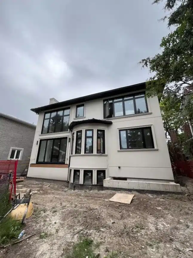 Full rear exterior view of the completed white stucco custom residential home featuring large windows, a central curved bay section, and a graded dirt yard under an overcast sky.