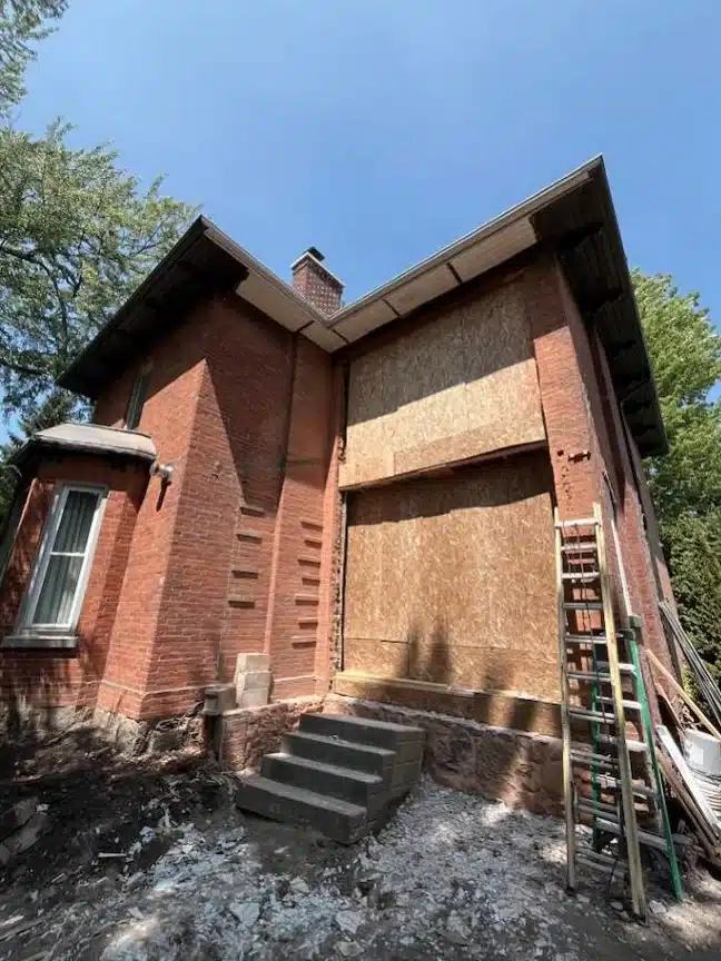 Exterior side view of a heritage red brick home with a large multi-storey structural wall opening securely boarded up with plywood panels during a major alteration.