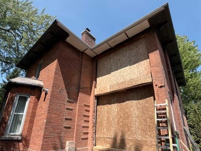 Exterior side view of a heritage red brick home with a large multi-storey structural wall opening securely boarded up with plywood panels during a major alteration.