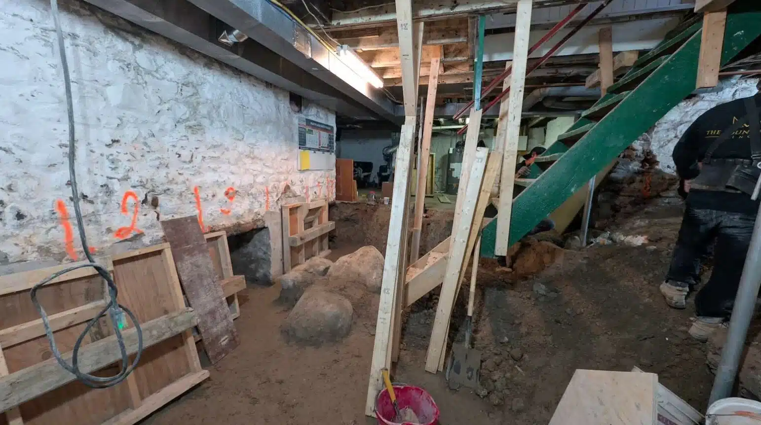 Interior structural framing view of a basement renovation with temporary vertical wood and steel shoring posts bracing the overhead joists during a foundation deep-lower project.