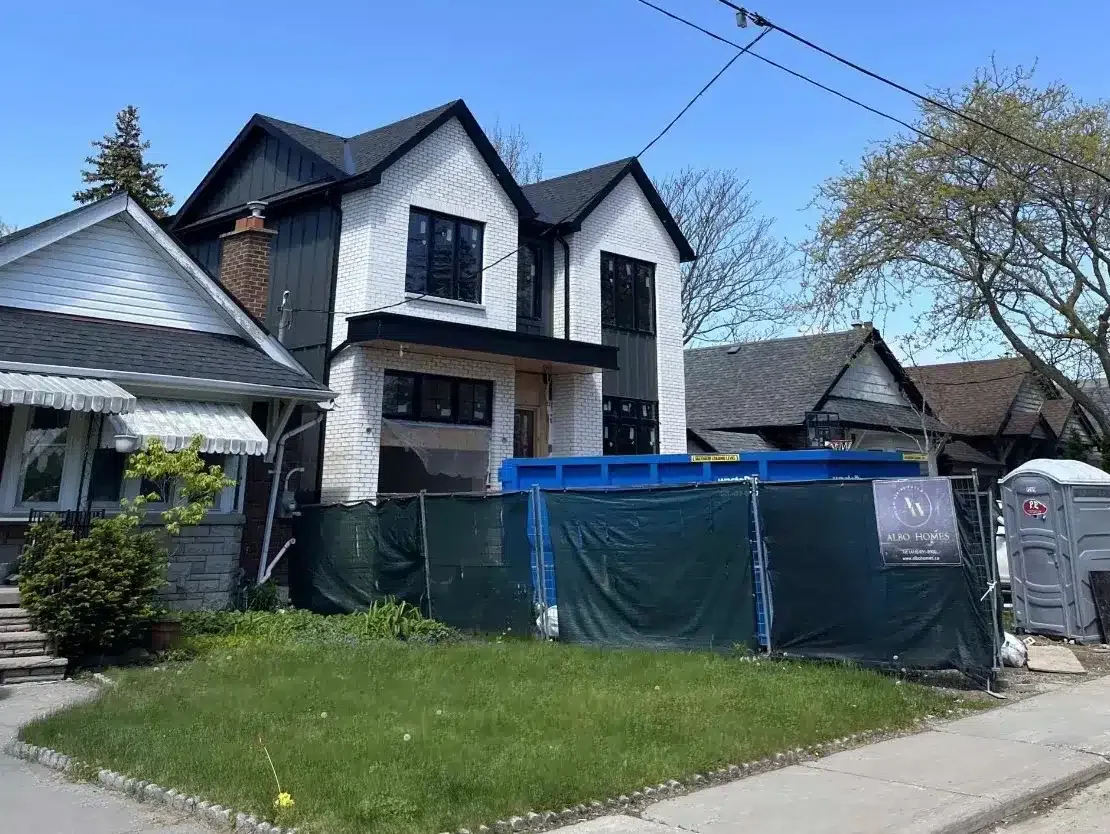 Completed exterior of a modern double gabled custom home with white brick veneer and black window trim behind a construction safety fence.